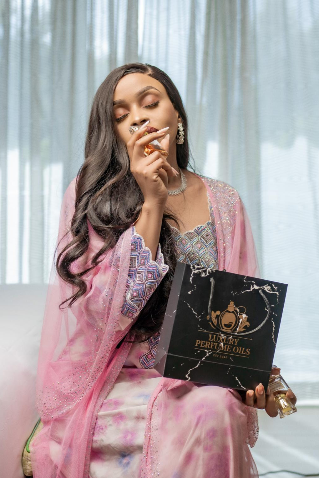 Woman in a pink outfit holding a black box with 'Perfume Oils' text, sitting on a couch.