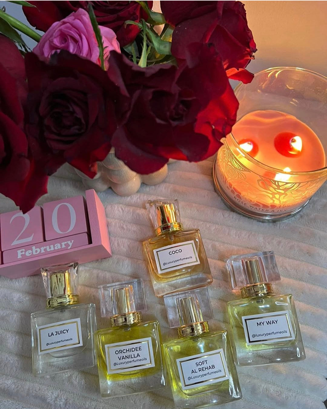 An assortment of @Luxuryperfumeoils glass bottles including scents like Orchidee Vanilla and Soft Al Rehab, styled with Valentine’s Day-themed decor including red roses and a lit "Rose Water & Ivy" candle.